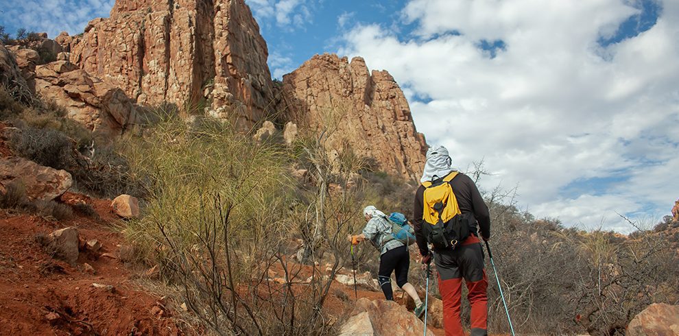 Two hikers walk up a rocky trail with trekking poles. Tall, reddish rock formations rise in the background under a partly cloudy sky, with dry shrubs along the path