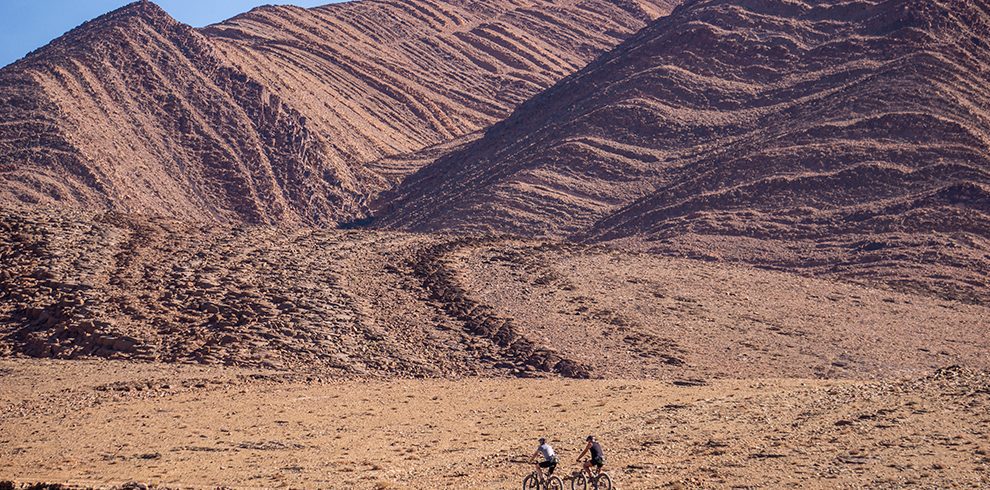 Two mountain bikers ride along a dry, rocky path in a vast desert landscape, with layered, reddish-brown hills rising behind them